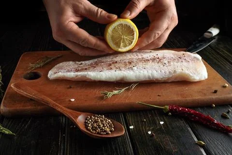 Hands squeeze fresh lemon over a fillet of fish resting on a wooden cutting.. Stock Photos