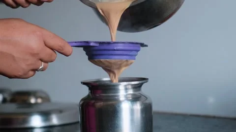 Hands straining hot Indian masala tea through a purple sieve into a steel pot Stock Footage 329975158