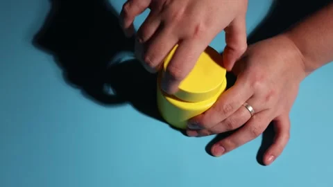 Hands twisting open a bright yellow container on a blue surface with dramatic Stock Footage 339177819