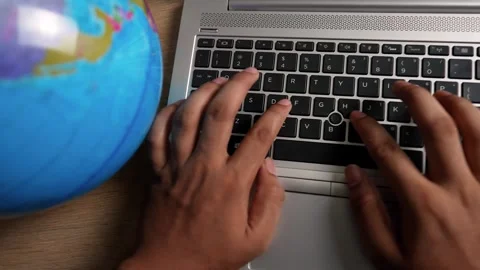 Hands typing on a laptop keyboard next to a globe on a wooden desk Stock Footage 317736772