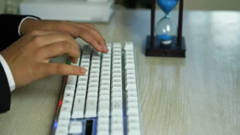 Hands typing on a white mechanical keyboard on a wood desk with an hourglas.. Видео 329177120