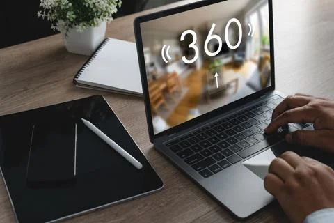 Hands using a laptop with a 360 virtual home tour on screen beside tablet a.. Stock Photos
