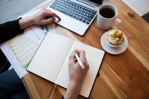 Hands using laptop and notebook with cup of tea and cake on wooden table in c Stock Photos