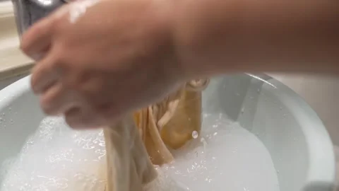 Hands Washing Beige Fabric in Soapy Water Basin, Close-up Shot Stockbeeldmateriaal 337414369