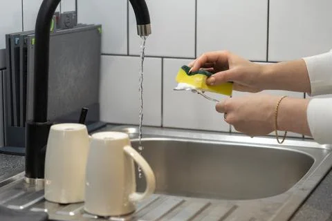 Hands washing a fork with a soapy yellow sponge under running water in a mo.. Stock Photos