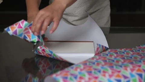 Hands Wrapping a Gift Box with Colorful Wrapping Paper on a Glass Surface Stock Footage 316839660