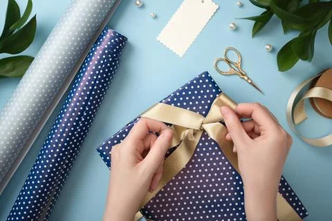 Hands wrapping a gift box with satin ribbon on blue background. Flat lay with Stock Photos