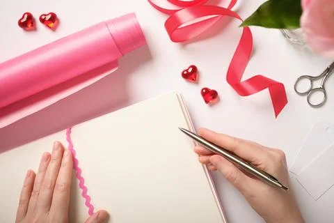 Hands writing in blank notebook surrounded by pink wrapping paper, red hearts Stock Photos