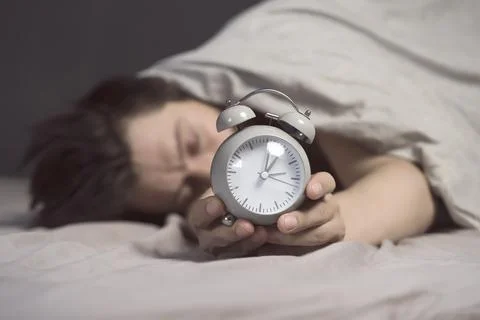 Hands of a young man from under the blankets hold an alarm clock in gray. Stock Photos