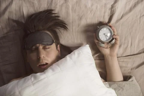 Hands of a young man from under the blankets hold an alarm clock in gray. Stock Photos