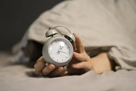 Hands of a young man from under the blankets hold an alarm clock in gray. Stock Photos