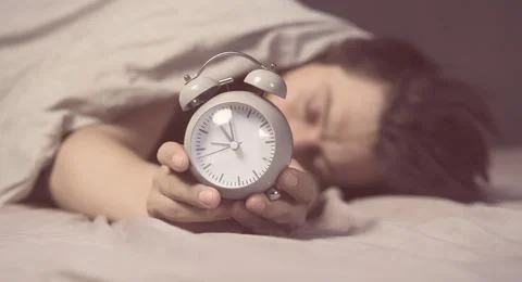 Hands of a young man from under the blankets hold an alarm clock in gray. Stock Photos