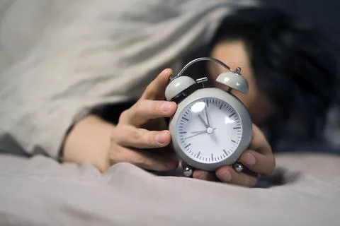 Hands of a young man from under the blankets hold a retro vintage alarm clock. Stock Photos