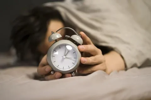 Hands of a young man from under the blankets hold a retro vintage alarm clock. Stock Photos