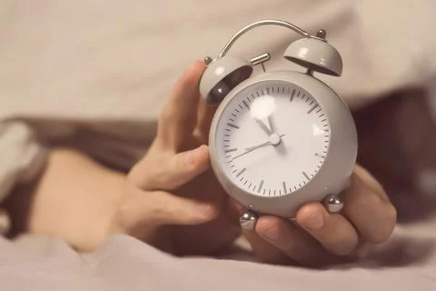 Hands of a young man from under the blankets hold a retro vintage alarm clock. Stock Photos