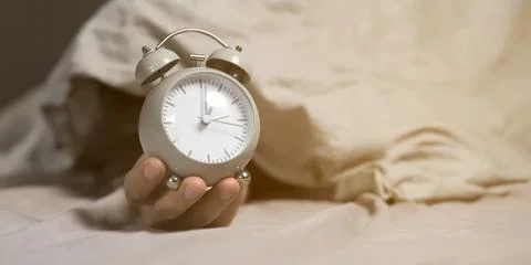 Hands of a young man from under the blankets hold an alarm clock in gray. Stock Photos