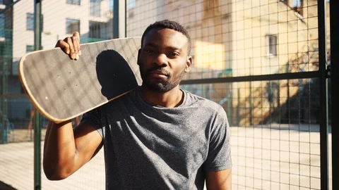 Handsome African guy in gray T-shirt holding black skateboard in hand, looking Stock Footage 92054577