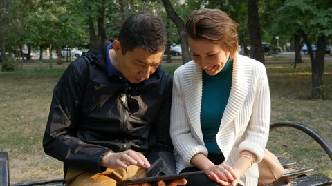 Handsome asian guy holding touch screen tablet with Caucasian girl in the park. Video stock 95598700