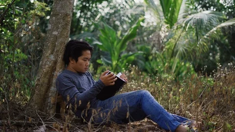 Handsome Asian young man sitting under tree, reading a book Stock Footage 93334283