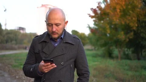 Handsome bald-headed man walking in the park and chatting on cellphone and Stock Footage 97116363