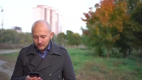 Handsome bald-headed man walking in the park and chatting on cellphone. Camera Stock Footage 97116594