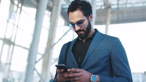 Handsome bearded Eastern businessman standing by the airport entrance, using his Video stock 76734877