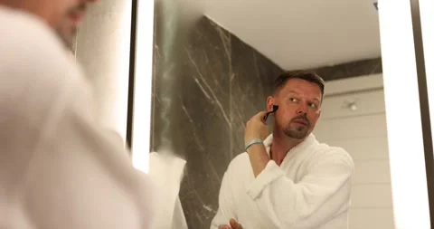 Handsome bearded guy shaves with disposable razor in bathroom and looks in Stock Footage 251739640