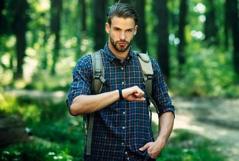 Handsome bearded man with backpack in checkered shirt looks at clock walk in  Stock Photos