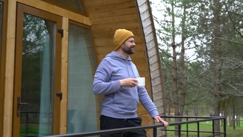 Handsome bearded man drinking hot tea in front of a modern wooden house Stock Footage 240955049