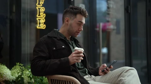 Handsome bearded man sitting outside cafe, enjoying cup of coffee and using Stock Footage 297624418