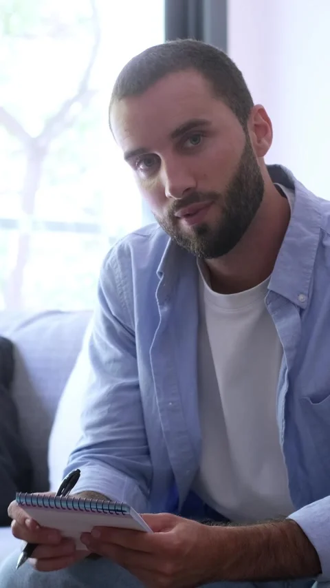 Handsome bearded man sitting on a sofa in a bright living room, attentively Stock Footage 345114377