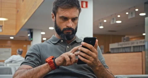 Handsome bearded man texting message on mobile phone while sitting at hospital 스톡 동영상 143478730