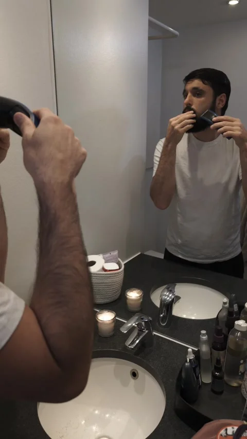 Handsome bearded man trimming beard with electric razor in bathroom mirror Video stock 335638065