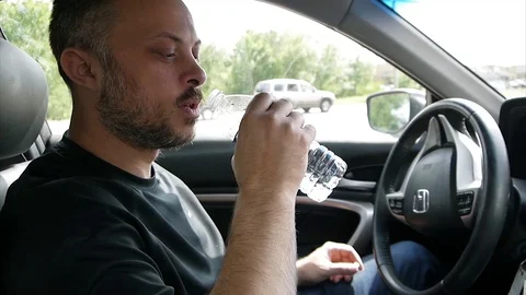Handsome bearded white man drinks water from a plastic bottle sitting in the car Stock Footage 77632527