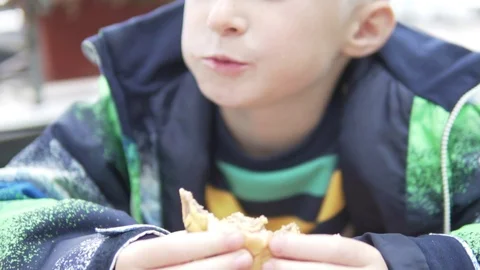 Handsome blond boy eagerly eats a cheeseburger on the street in autumn Stock Footage 116098762