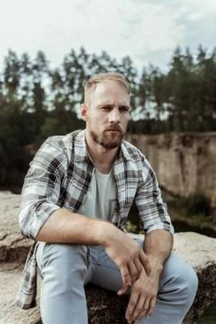 Handsome blonde-haired model posing for photo while sitting on rock near canyon Stock Photos