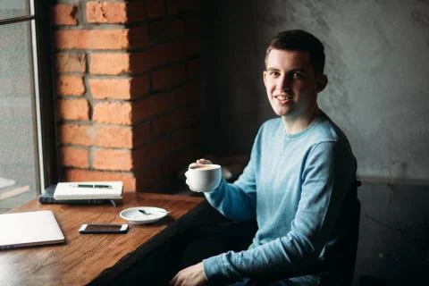 Handsome boy drinking coffee in cafe. He sits near the window with laptop and Stock Photos