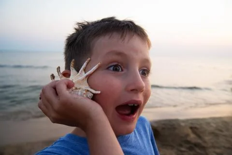 Handsome boy with a surprised expression on his face listens to a seashell. T Stock Photos
