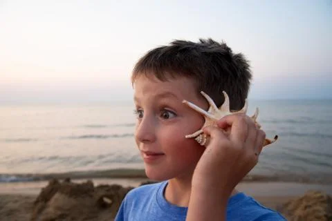 Handsome boy with a surprised expression on his face listens to a seashell. T Stock Photos