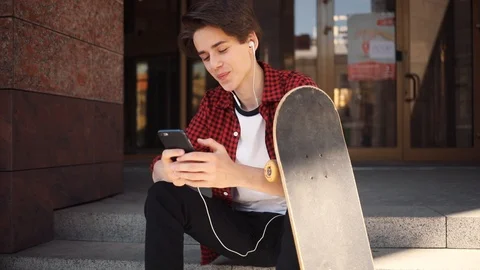 Handsome boy using mobile phone with headphones sitting keeps skate guy business Stock Footage 92051579
