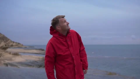 Handsome brunette man in red jacket looking at overcast sky on a cliff by the Stock Footage 274050985