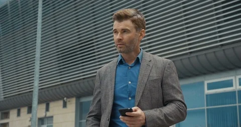 Handsome Businessman Chatting on his Smartphone While Exiting the Big Building Stock Footage 114758564