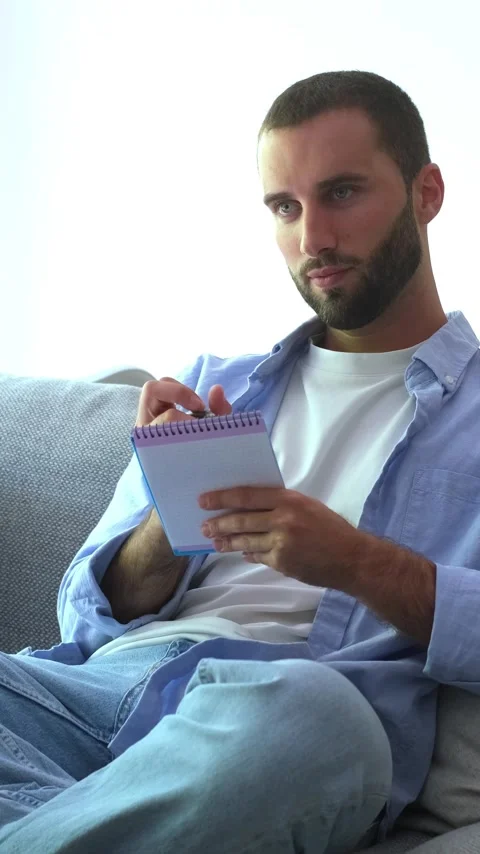 Handsome caucasian man with a beard sitting on a couch, thoughtfully writing Vídeos de archivo 344782195