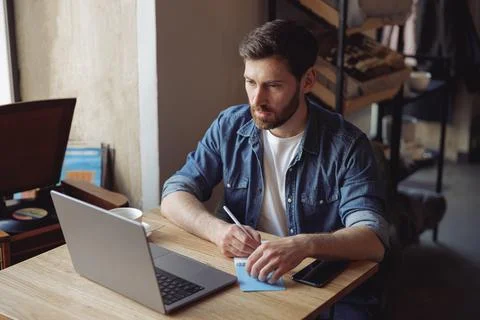 Handsome Caucasian man taking notes in notebook and sitting in cafe at laptop 스톡 사진
