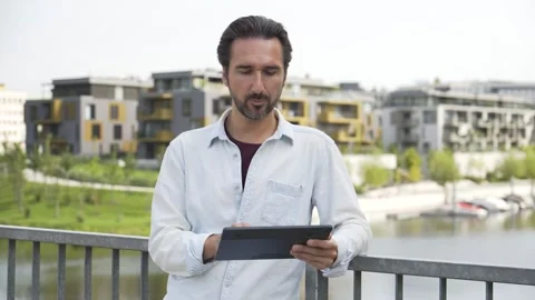 A handsome Caucasian man talks to the camera and works on a tablet - a river and Stock Footage 277905968