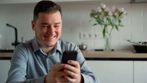 Handsome caucasian man using smartphone while sitting on a chair in kitchen Stock Footage 153913633
