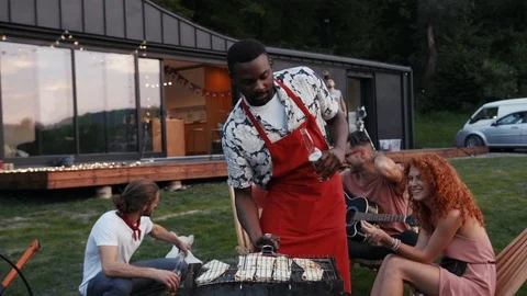 Handsome chef in red apron cooking outdoors on grill, drinking beer, smiles Stock Footage 91929542