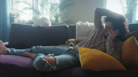 A handsome curly-haired man on a couch surrounded by pets talking on the phone. Stock Footage 193979637