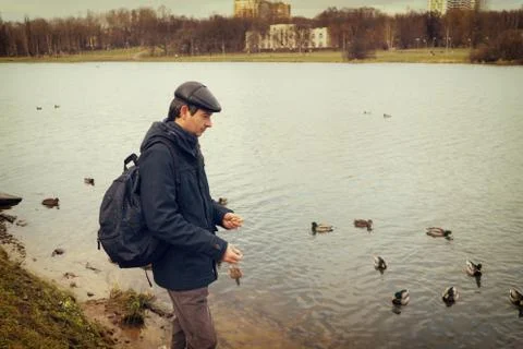 Handsome dark-haired middle-aged man in jacket, cap and backpack feeds ducks  Stock Photos