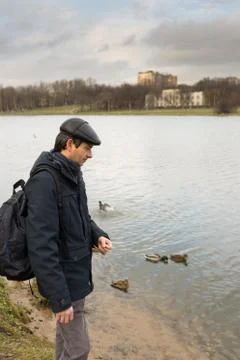 Handsome dark-haired middle-aged man in jacket, cap and backpack feeds ducks  Stock Photos
